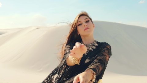 A Beautiful young woman fashion model posing in a desert. Adult Girl dancing with her hands in long silk black dress. The brunette hair flies in wind. background nature dune sand, blue sky