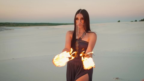 Beautiful young woman with fire in her hands posing at camera. Pretty Face hides gold veil mask chain. United Arab Emirates desert background. White sand at sunset. Mysterious oriental seductive Queen
