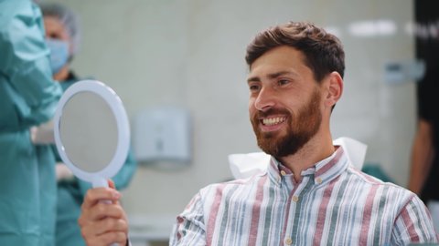 Handsome cheerful young male patient smiling into mirror looking at healthy white teeth enjoying dental healthcare facilities in modern dentist office.