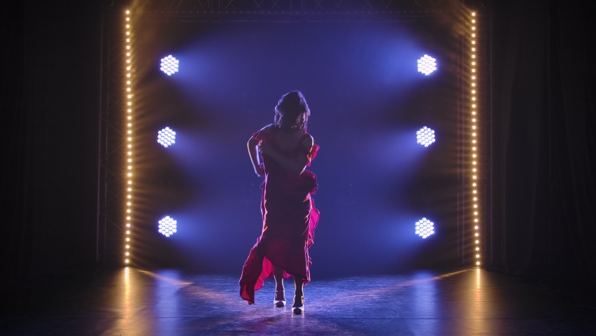 Silhouette of a slender dancer dancing passionate Argentine flamenco in a dark studio against the backdrop of lights. The graceful movements of a dancer in slow motion.