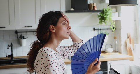 Overheated millennial 35s female standing in domestic kitchen using handheld blue waver, waves herself with hand fan suffers from hot weather indoor. Period, hormonal imbalance unhealthy woman concept
