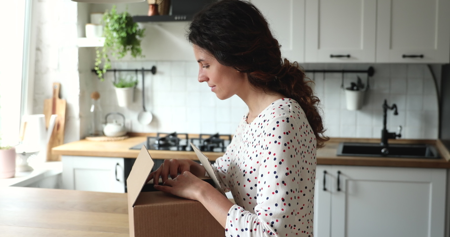 Woman received gift from foreign friend by mail, stands in kitchen opens parcel box looks inside examine carefully ordered high quality goods feels glad. Safe secure express delivery services concept | Shutterstock HD Video #1060760167