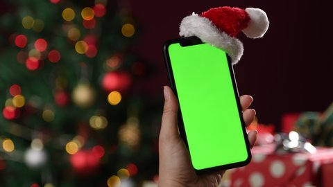 Hand holding the black smartphone with green screen and Santa Claus' hat on top on Christmas background
