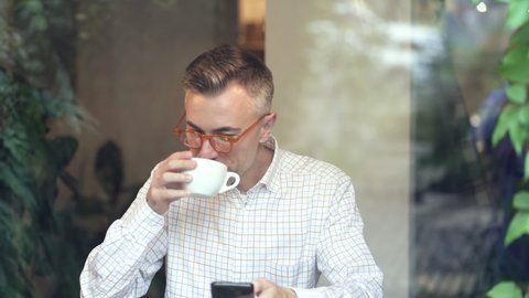 Happy man drinking coffe from white cup and holding mobile phone. Looking at the mobile phone. Slowmotion.