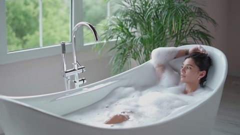 Relaxed woman in bath in slow motion. Sexy girl taking bathtub with bubbles and foam in slow motion. Top view of pretty lady enjoying in luxury bathroom