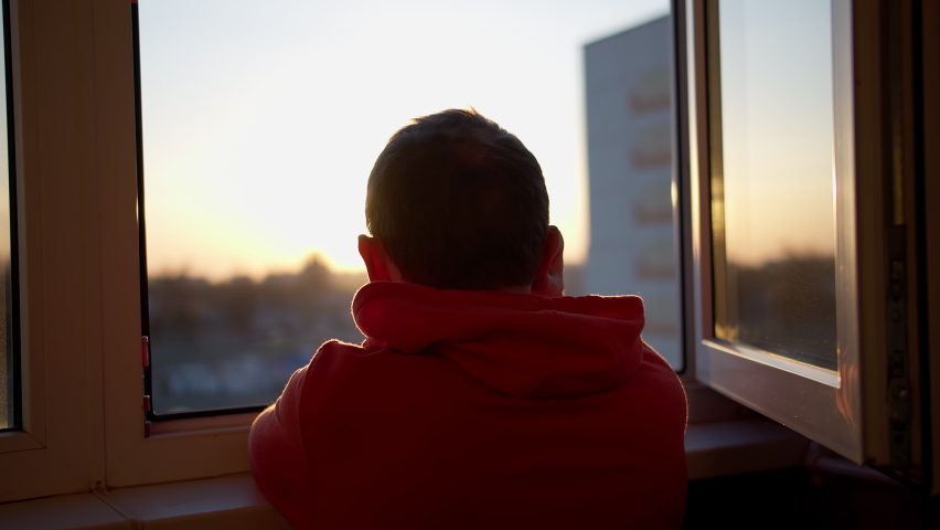 Man in a sweatshirt looks at the sunset while standing in an open window, camera movement