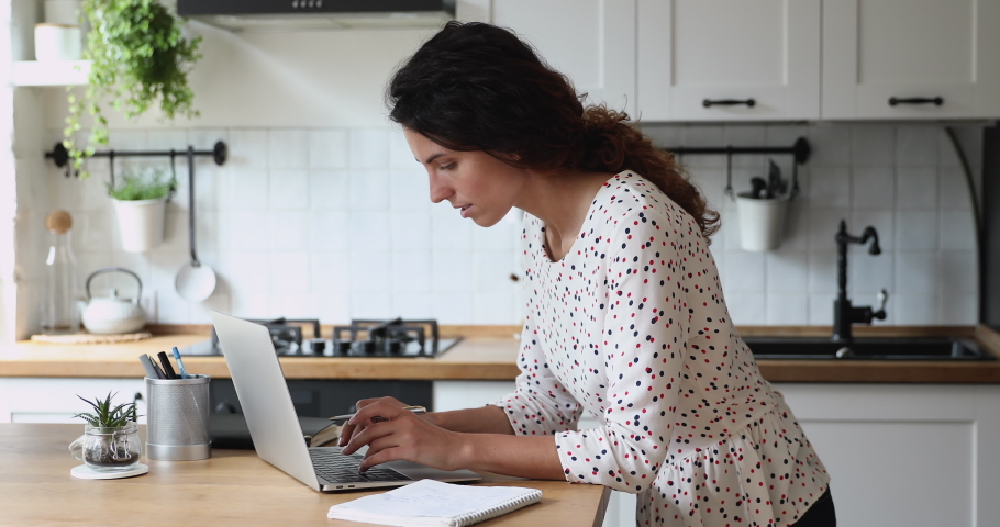 Attractive 35s freelance woman standing in home kitchen use laptop search information on internet write down it in copybook do telework, she busy with self-education professional development concept Royalty-Free Stock Footage #1060767088
