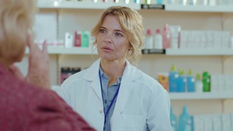 Blond woman pharmacist serving a customer in a drugstore. Conversation pharmaceutical client. Seller commercial health care buyer uniform. Slow motion