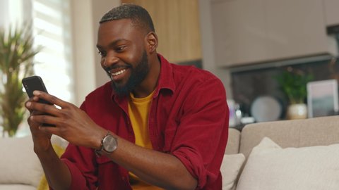 Wonder happy young african american man use phone smiling read good news sit on sofa in home. Communication in social media online relax work app. Handsome guy. Internet. Slow motion