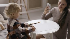 Young mother takes pictures and video of her daughter on smartphone, who writes a letter to Santa Claus and draws. Christmas tree background.