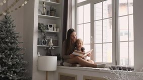 Mother and daughter having fun on Christmas morning. Young mom reading a book with her toddler daughter by the window. - Powered by Shutterstock - Get 15% off with code: PIKWIZARD15