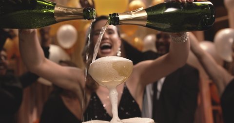 Young woman filling champagne into a tower of glasses with friends dancing around at gala night party. Group of multiethnic people having a great time at new years party.
