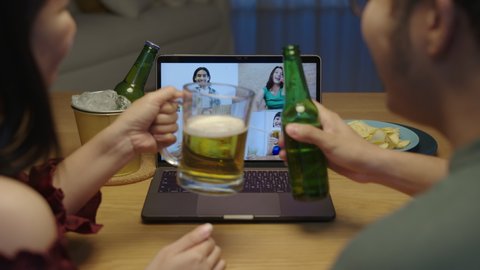 Zoom out : Young happy asian couple enjoy relax night party event online celebration festive with friends at home clinking beer with glass and bottle toasting drinking via video call.