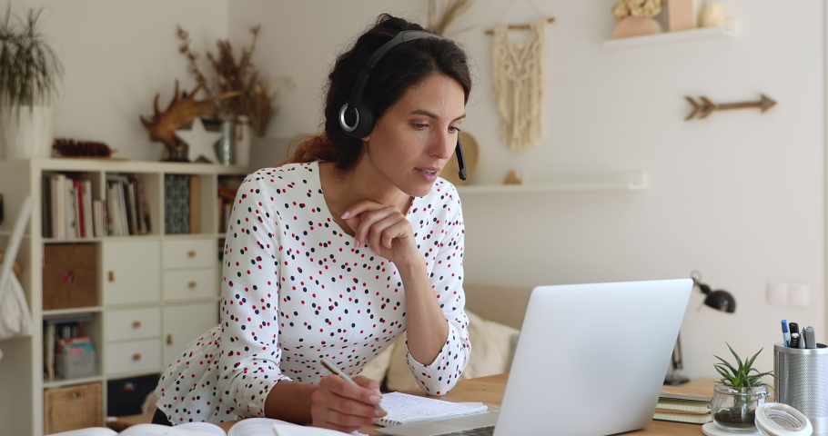 35s woman lean at table wear headset looks at laptop screen writing down received information, listen e-teacher, gain new knowledge by videocall. Take part in webinar conducted over internet concept Royalty-Free Stock Footage #1060771897
