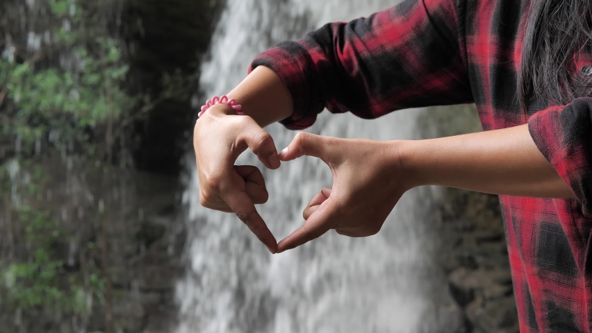 A woman uses her hand to make a heart symbol with a beautiful waterfall, the woman is happy with the waterfall.