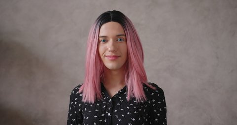 Elegant young transwoman wearing stylish black dot dress with bright pink wig smiles to camera standing on beige background