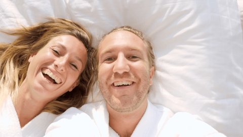 SLOW MOTION: Happy young confident couple taking selfies on the bed wearing a bathrobe. Young couple taking self portraits in a hotel room on a comfy bed