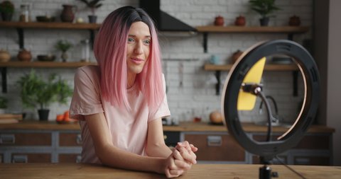 Pretty transwoman with pink wig shoots new video with smartphone and ring light sitting at table in decorated kitchen at home