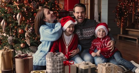 Beautiful family with little children celebrate Christmas at modern ornate home. Married couple their pretty kids hugging seated near gift boxes and twinkle lights festive fir laughing feels happy