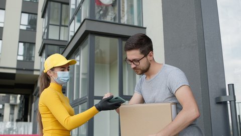 Man Opens The Door. Young Girl Delivery Service Worker Brings Customer Box. Guy Takes The Box And Puts His Signature On The Girls Phone. She Is Wearing Gloves And Mask To Protect Against Viral