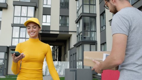 Young Girl With Delivery Service Gives Box To The Customer. Pretty Girl In Yellow Cap With Long Hair. Handsome Guy With Glasses Takes Box And Smiles. He Puts Signature On The Girls Phone. Meeting On