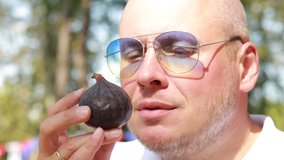 Video, an emotional adult man in a white shirt and sunglasses eats fresh fig fruit in a public park. On a sunny day in summer.