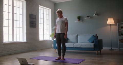 Mature caucasian woman is exercising at home, watching an online video on her laptop and stretching, keeping herself fit 4k footage
