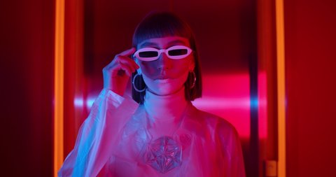 Portrait of female young clubber in futuristic outfit taking off sunglasses while standing in hall with neon lights. Woman with make up wearing trendy clothes looking to camera .