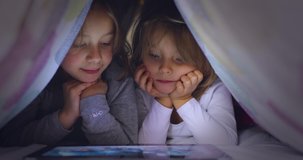 Child blonde caucasian girl with friend or sister watching movie or animation on tablet app under bed blanket at night. Modern technology childhood kid use at home. 4k video