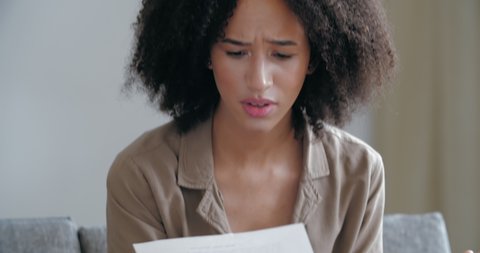 Young mixed race woman sits at home on couch with notice letter in her hands, reads the information text about dismissal from work, feels anger frustrated, sadly excitedly holds head with her arms