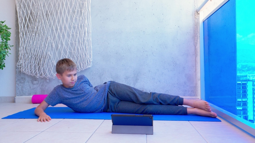 Boy doing sport exercises while watching video lesson online on tablet computer on balcony. Sport, healhty lifestyle, active leisure at home