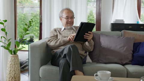 Candid of old asian retired man using tablet computer to transfer money online or financial payment and read text, smile or laugh with happy and positive. Senior asian with digital technology concept