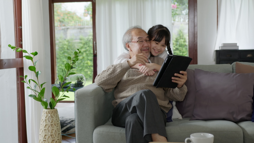Candid of old senior asian grandparent play and watch with kid grandchildren with technology on computer tablet at home in bonding relationship in family. Young girl hug older man from back.