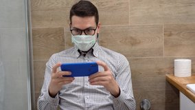 Happy young Caucasian handsome brunette man in gray plaid shirt in protective face mask wear black glasses, sit in toilet, hold blue smartphone, plays in game on phone, close up on background of tiles