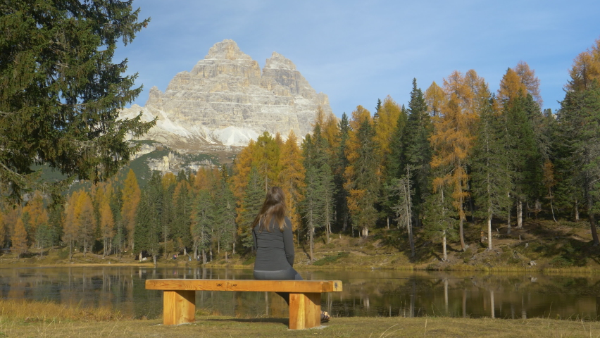 LOW ANGLE, COPY SPACE: Unrecognizable young woman sits on an empty wooden bench and observes the breathtaking fall colored mountain and lake landscape. Hiker enjoying the scenic views of lake Antorno.