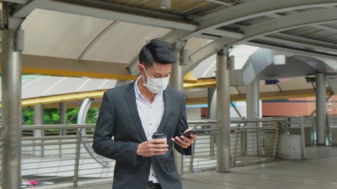 Asian young businessman walking up on stair going to work. The working man holding coffee cup and mobile phone walking to office wearing suit and face mask for prevent COVID infection.