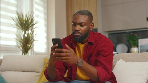 Focused serious young african american man use phone look around sit on sofa in home. Communication in social media online relax work app. Handsome serious guy. Internet. Slow motion
