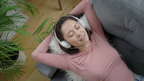 Young mixed race woman relaxing on comfortable sofa with closed eyes wearing headphones, asian girl enjoys listening chill music and audio sound relaxing at home. Top view in slow motion