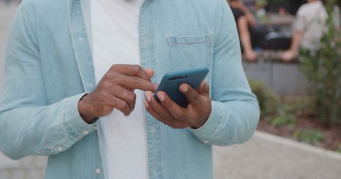 Unknown stylish dark-skinned man in casual clothes standing outdoors near cafe and browsing phone apps,close up