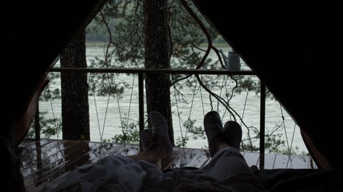 Couple in White Bathrobes and Slippers Lying in Bed in Glamping Tent on Rainy Day. Beautiful Scenic View of Mountain River is on the Background. Slow Motion. Camping, Traveling, Tourism