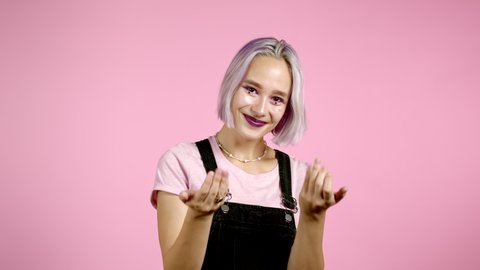 Beautiful hipster woman showing - Hey you, come here.Girl in overall ask join her, beckons with inviting hand hugs gesture.Lady is looking playful flirtatious, inviting to come.Pink studio background.