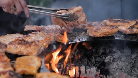 Chef holding tongs and grilling fresh juicy meat steaks on round brazier with hot flame at summer local food market - close up view, slow motion. Outdoor cooking, gastronomy, street food concept