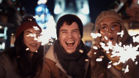 Three happy friends with sparklers call their relatives via video link on new year's eve to the sound of chimes in the middle of the street in the lights of the Christmas market against the background