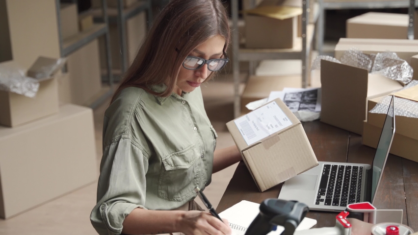 Young female seller or online store worker holding parcel box checking ecommerce post shipping online retail e commerce store order fulfillment using laptop in dropshipping delivery service warehouse. | Shutterstock HD Video #1060827886