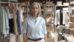 Confident happy mature older 60s woman retail seller, entrepreneur, clothing store small business owner, supervisor looking at camera standing arms crossed in delivery shipping warehouse, portrait.