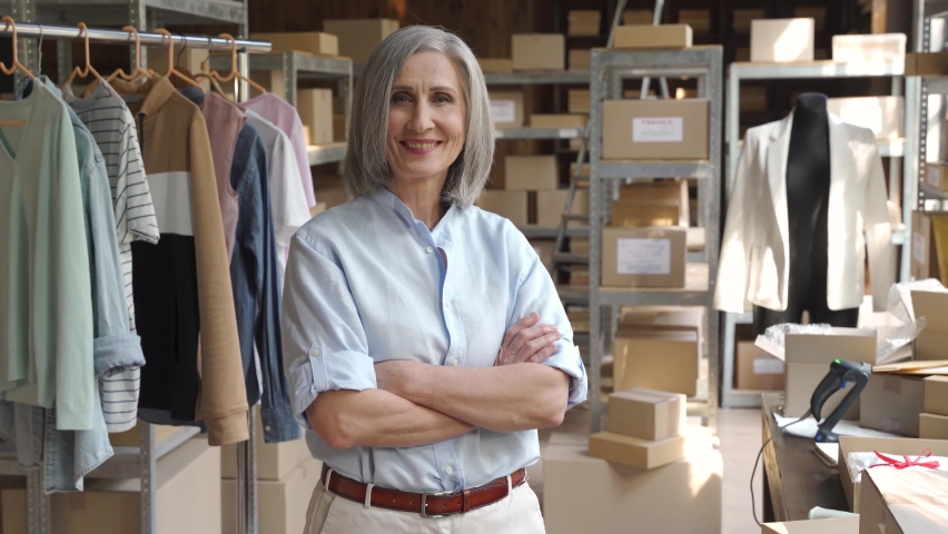 Confident happy mature older 60s woman retail seller, entrepreneur, clothing store small business owner, supervisor looking at camera standing arms crossed in delivery shipping warehouse, portrait. Royalty-Free Stock Footage #1060830634