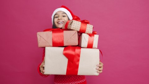 Funny indian latin preteen girl wears santa hat holding many gifts dropping boxes falling down standing on red background. Merry Christmas presents shopping sale, Happy 2021 New Year celebration.