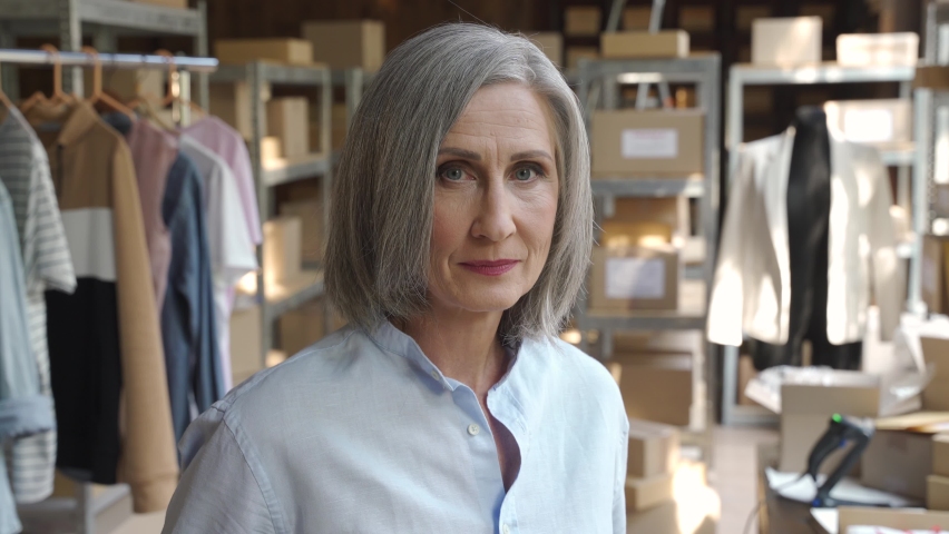Mature older 60s woman fashion seller, entrepreneur, clothing store dropshipping small business owner looking at camera in postal delivery shipping warehouse stock office, close up headshot portrait. Royalty-Free Stock Footage #1060830643