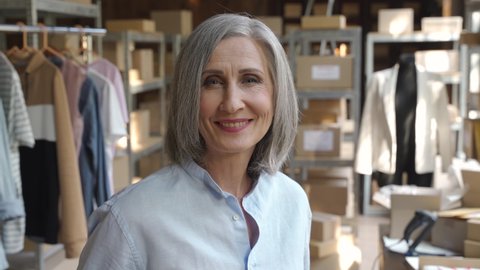 Mature older 60s woman fashion seller, entrepreneur, clothing store dropshipping small business owner looking at camera in postal delivery shipping warehouse stock office, close up headshot portrait.