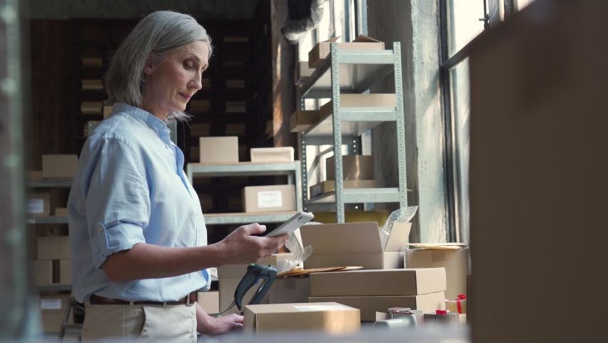 Female mature senior small business owner using mobile app checking parcel box. Warehouse worker, seller holding phone scanning retail dropshipping package postal parcel on cell preparing ship order. Royalty-Free Stock Footage #1060830652